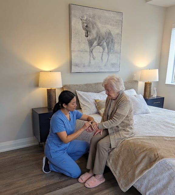 A woman in blue scrubs is holding the hand of an elderly woman in a beige outfit.