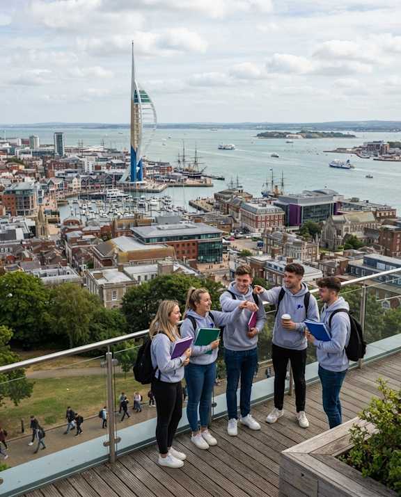 Four students are standing on a balcony overlooking a cityscape with a body of water and boats.