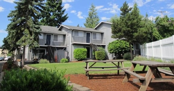 A wooden picnic table is in front of a building with a white picket fence.
