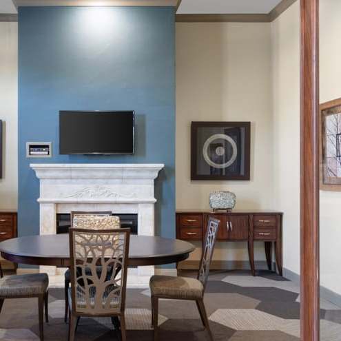 The amazing resident lounge with a cozy fireplace with a TV over it on a blue accent wall, two small dressers, wall art, and a circular dining table with chairs with the backs made into tree patterns at the Landing at Mansfield Apartments. 
