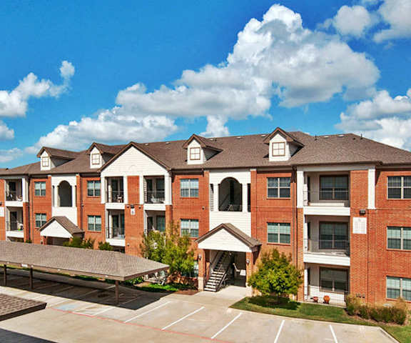 The exterior of the red brick apartment buildings with trees and patios in front, and the covered parking lot for residents at the Landing at Mansfield Apartments. 