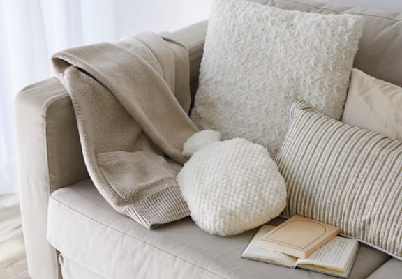A beige couch with a white pillow, a white knitted pillow, a book, and a beige blanket.