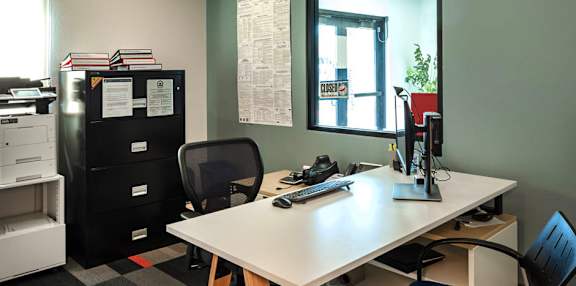 A black fridge sits next to a desk with a computer on it.