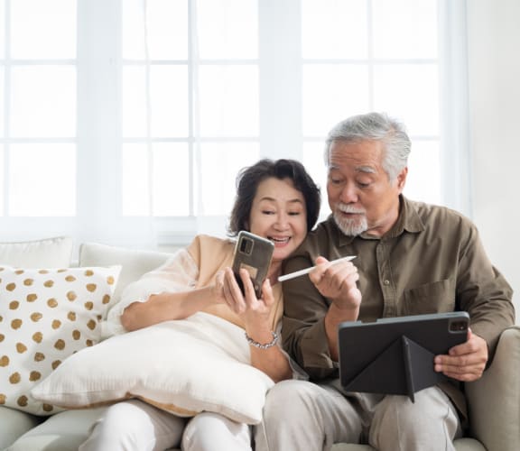 an older couple sitting on a couch looking at their cell phones and a tablet