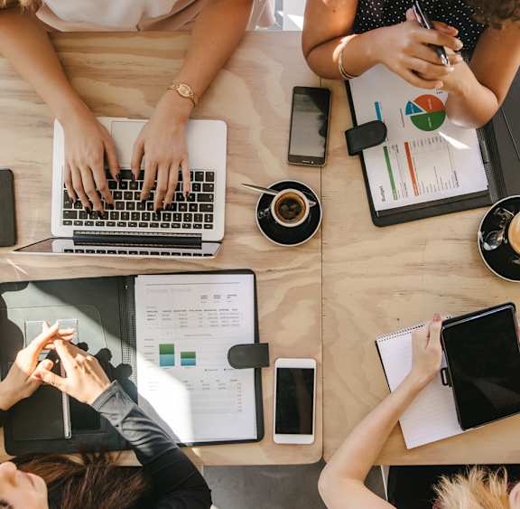 A group of people are gathered around a table with laptops, tablets, and papers, appearing to be in a meeting or collaborative work session.