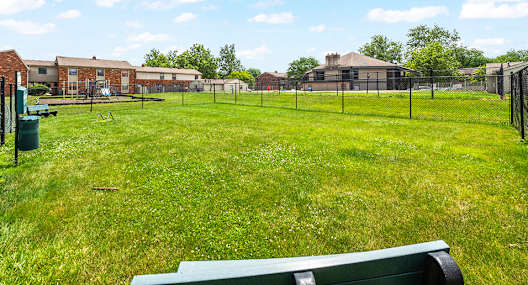 A green bench is in the foreground of a grassy field with a fence and buildings in the background.