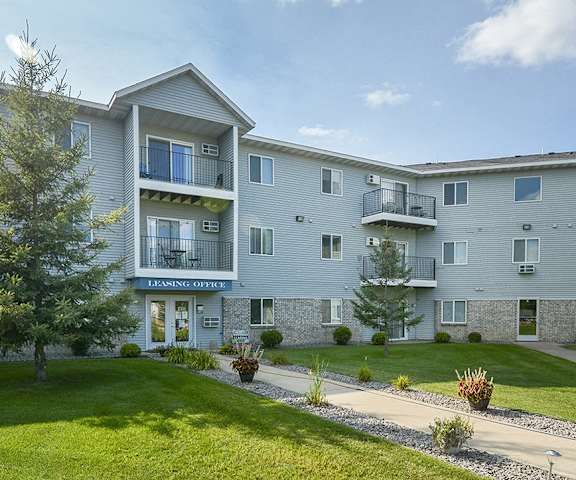 The exterior of the leasing office in the light blue apartment building with a pathway surrounded by grass, bushes, and trees at Quarry Commons Apartments. 