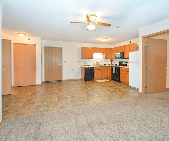 A view of the empty carpeted living room with a window and the kitchen with white appliances and wooden large cabinets at Quarry Commons Apartments. 