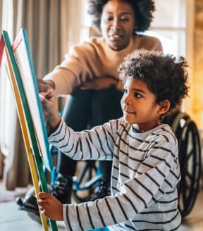 a boy in a wheelchair paints with a paintbrush with his mother in the background