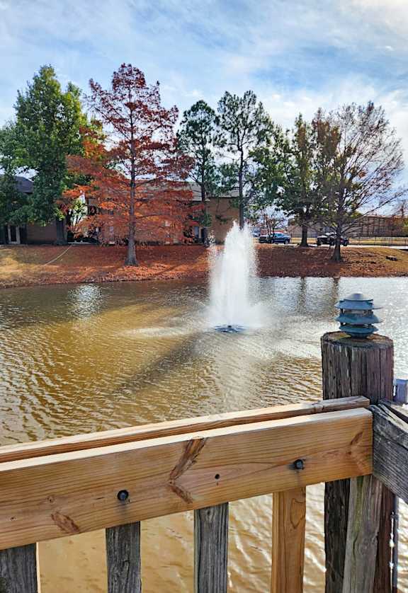 A fountain in the middle of a pond with a wooden fence in the foreground.