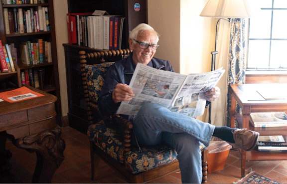 An elderly man is reading a newspaper while sitting in a chair.