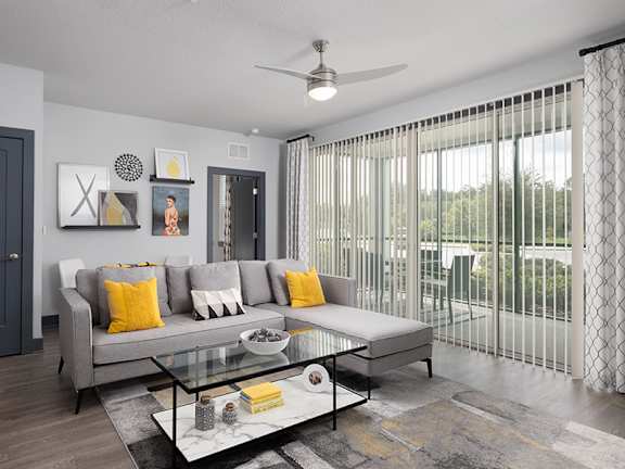 A modern living room with a grey sofa, a glass coffee table, and a ceiling fan.