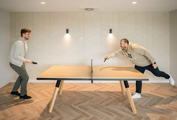 Two men playing ping pong in a room with wooden flooring.
