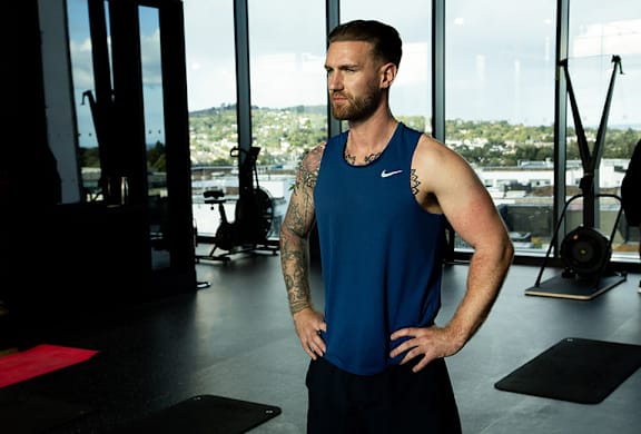 A man with tattoos stands in a gym wearing a blue Nike tank top.
