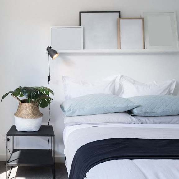 A bedroom with a bed, a plant on a table, and picture frames on a shelf.