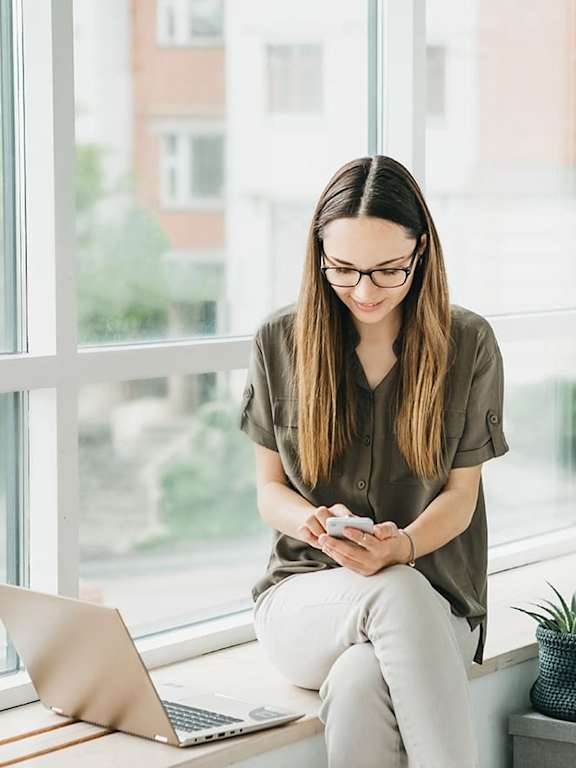 A woman is sitting by a window with a laptop and a smartphone in her hands.
