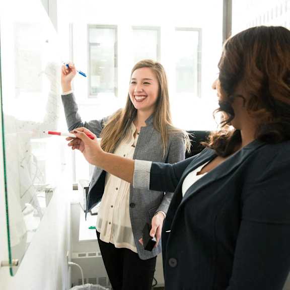 two women writing on a whiteboard with a marker