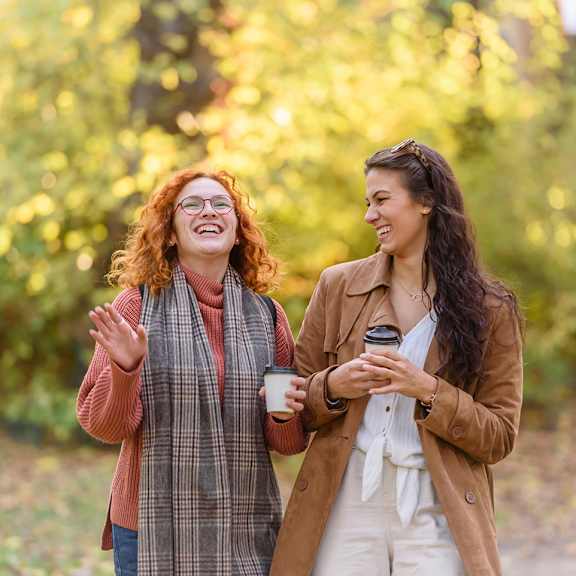 Two women are standing outside, one holding a coffee cup and the other holding a phone.