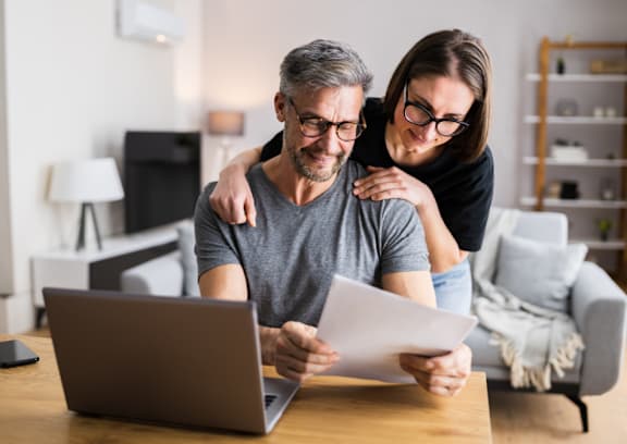 A man and woman are looking at a document together while sitting at a table with a laptop.