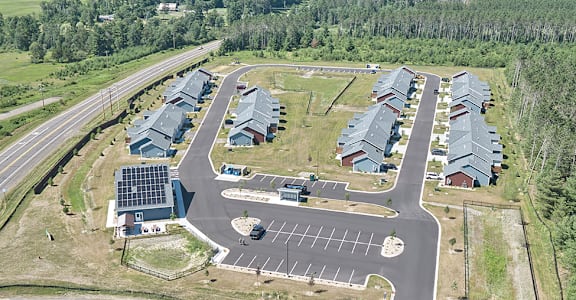 A road with a car driving down it and a parking lot with a building in the background.
