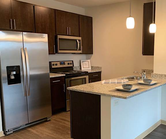 A large, modern kitchen with stainless steel appliances, bar counter with barstools, kitchen utensils, marble countertops with lots of room, two places set on the kitchen bar, and large wooden cabinets at Royster Commons Apartment Homes. 