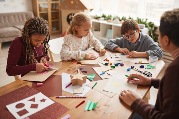Three children are sitting at a table working on art projects.
