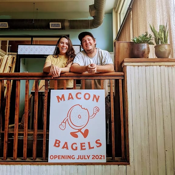 Two people are standing behind a sign that says Macon Bagels.