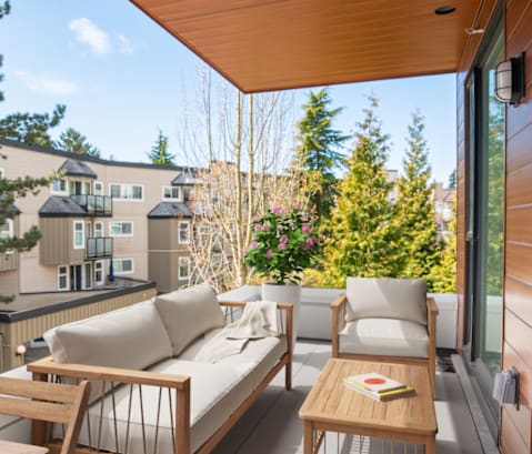 A wooden sofa and chair are on a balcony with a view of a building and trees.