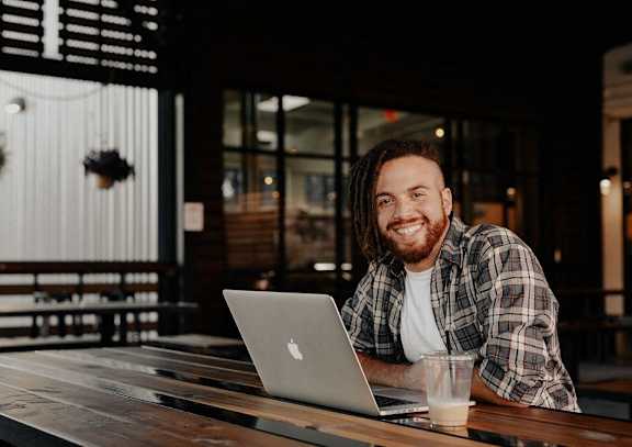 A man with a beard and dreadlocks is smiling while working on his laptop at a wooden table.