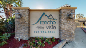 Apartments for Rent in Ontario CA - Rancho Vista - Front Exterior With Decorative Flowers, Trees, Lamps, and Apartment Logo