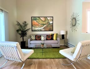 2 bedroom model living room with a tan couch, 2 white accent chairs, glass coffee table, green accent rug, plants, lamp and wall decorations