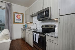 Half kitchen with wood textured gray cabinets, granite countertop and stainless steel appliances, open to living room
