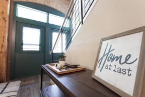 Entryway of unit with iron staircase leading up and black double doors with glass inserts opposite the stairs. Sign on console table by staircase reads "Home at last."