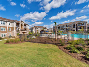 Daytime outdoor view of the gated pool surrounded by bushes, grass, and apartment complexes