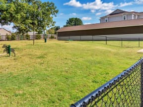Fenced-in dog park with a tall lamp, grass, and agility equipment near apartment parking garages