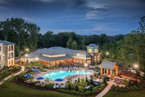 Aerial night view of lit pool, sundeck, outdoor lounge, and clubhouse with surrounding apartments