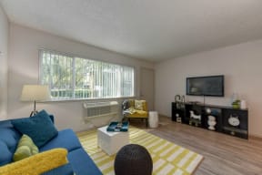 Living Room with Hardwood Inspired Floors, Yellow/White Rug, Large Window with Blinds, Blue Sofa and Flat Screen Television at Pinecrest Apartments, Davis ,95616