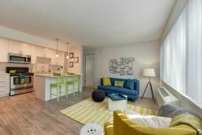 Living Room with Hardwood Inspired Floors, Yellow/White Rug, Large Window with Blinds, Blue Sofa and View of Kitchen at Pinecrest Apartments, Davis, CA, 95616