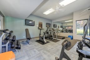 Community Fitness Center.  The Room has a light blue accent wall and contains a weight system, elliptical machines, treadmill, bench and free weights.