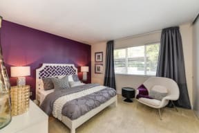 Master bedroom in the model home. Queen size bed with two nightstands. Dark Purple accent wall. There is a light colored lounge chair off to the corner.