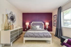 Master bedroom in the model home. Queen size bed with two nightstands both with white lamps on them. There is a long dresser along one wall.   There is a dark Purple accent wall