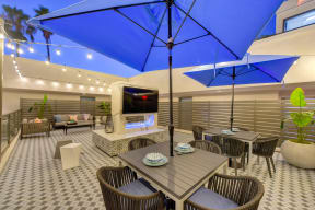 Outdoor rooftop lounge area at dusk.  There are bright blur patio umbrellas with tables, chairs and fireplace with flat screen TV above.