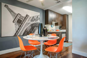 Model Dining area with table and 4 orange chairs.  View of the kitchen area  in the distance.  Hardwood inspired flooring through out the dining and kitchen area.
