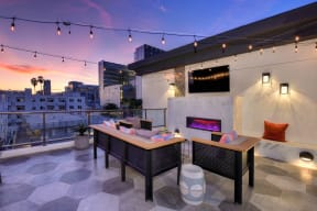 Roof top lounge area at sunset.  The sky is a beautiful blue and orange color. Lounge area comes with couches, chairs, fire pit and flat screen TV on wall. 