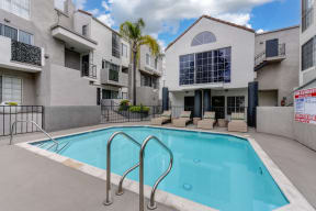 Community Swimming pool located in the middle of the apartment community with apartment buildings overlooking the pool deck.  There are wicker lounge chairs present.