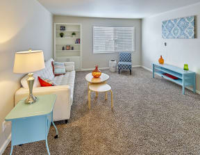 Large carpeted living space with window and staged with a couch, side tables, coffee table, chair and art.