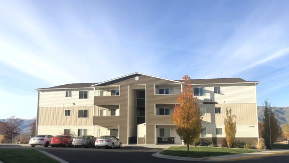 Exterior of Canyon View Apartments with mountains and blue skies behind, parked cars to the left, and landscaping on the right.