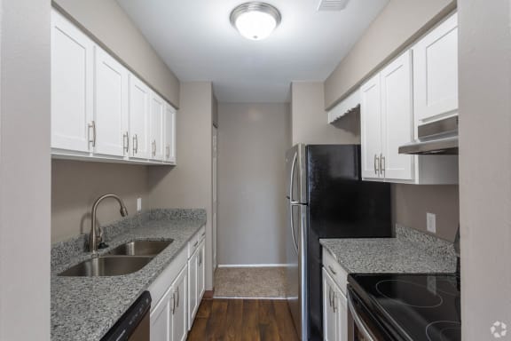 Galley-style  kitchen with granite speckled grey/white counters, white cabinets with pull handles on both lower and upper, hardwood-like flooring and stainless appliances