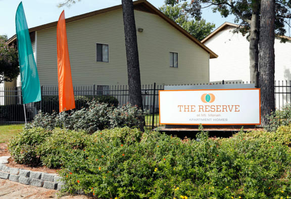 Monument Sign with The Reserve at Mt. Moriah in teal and orange surrounded by green bushes. Teal and orange sign to the left of sign.  Buildings in background.