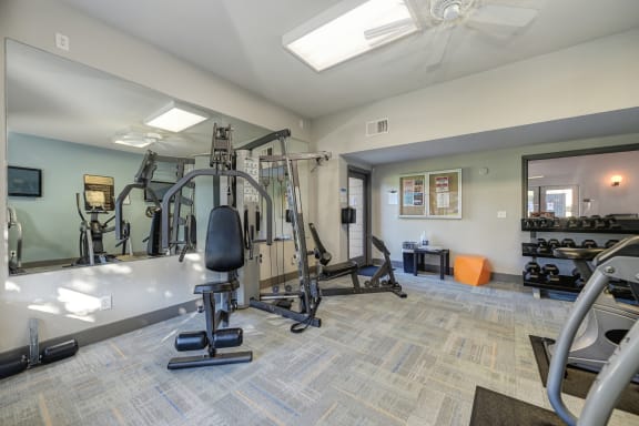 Community Fitness Center.  The Room has a light blue accent wall and contains a weight system, elliptical machines, bench and free weights.